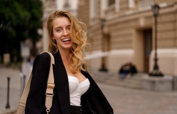 Beautiful smiling woman in elegant black suit posing outdoor in old european city. Blond wavy hairs, perfet skin.