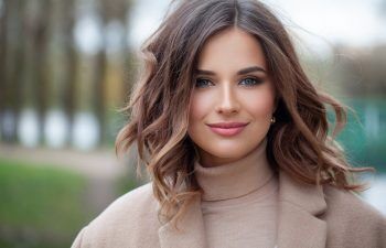A woman with wavy brown hair and blue eyes is wearing a beige turtleneck and coat, standing outdoors with a blurred background of trees and greenery.