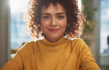 A person with curly hair and a yellow turtleneck sweater smiles while sitting indoors with sunlight in the background.